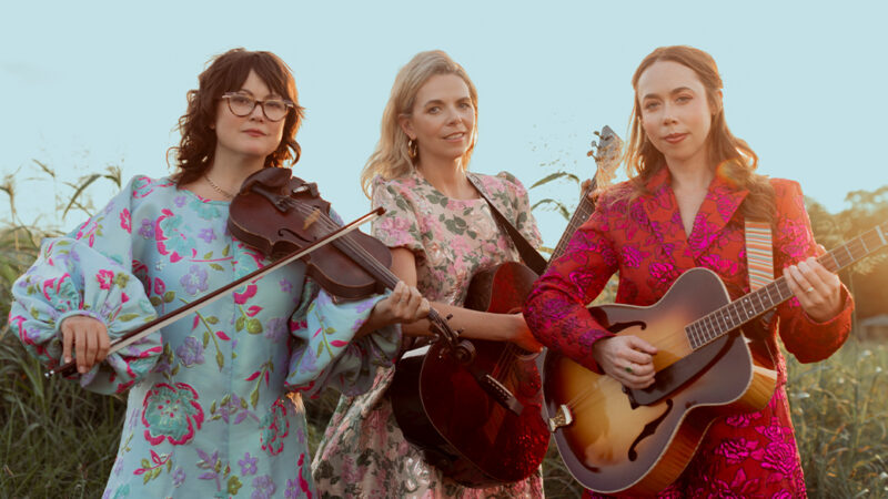 one brunette woman with long hair wearing a light blue dress with red flowers holding a violin. one woman with blonde long hair holding a guitar and wearing a pink floral dress. one woman with blonde long hair wearing a red dress holding a guitar.