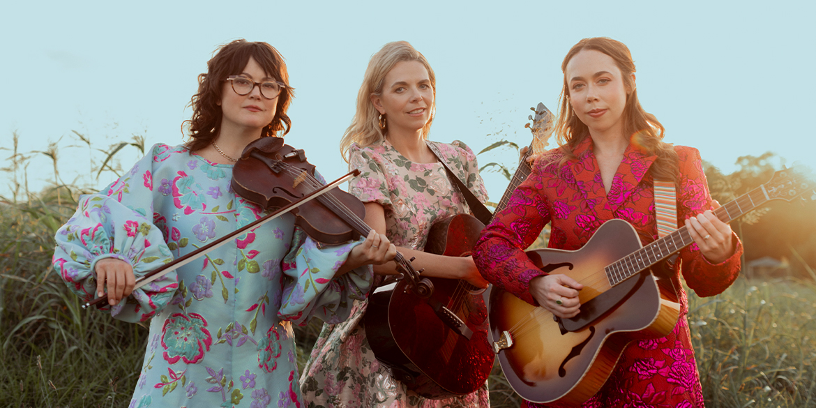 one brunette woman with long hair wearing a light blue dress with red flowers holding a violin. one woman with blonde long hair holding a guitar and wearing a pink floral dress. one woman with blonde long hair wearing a red dress holding a guitar.
