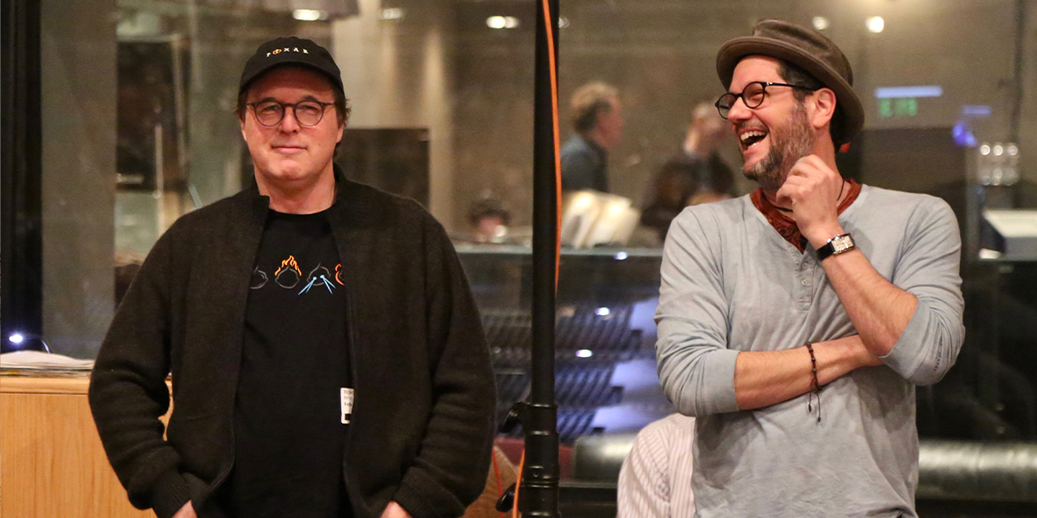 A side-by-side photo of two men in a dimly lit indoor setting, likely a recording studio. On the left, a man with gray hair and glasses wears a black baseball cap, a black graphic t-shirt, and a dark cardigan, standing with his hands in his pockets and a slight smile. On the right, a man with a beard and glasses laughs heartily, wearing a brown fedora-style hat, a gray long-sleeve henley, a red bandana around his neck, and a watch and bracelet on his wrist. A glass partition divides the two halves of the image, with other people and equipment visible in the background.